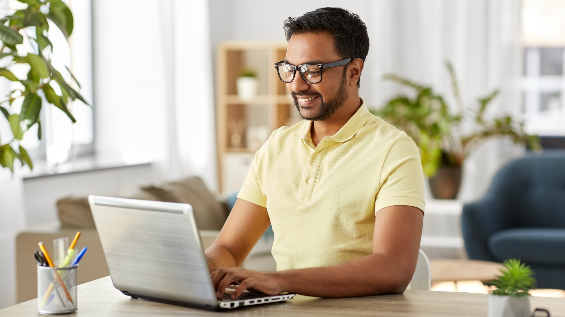 Man in yellow polo shirt smiling while sitting at a table and working on his laptop.