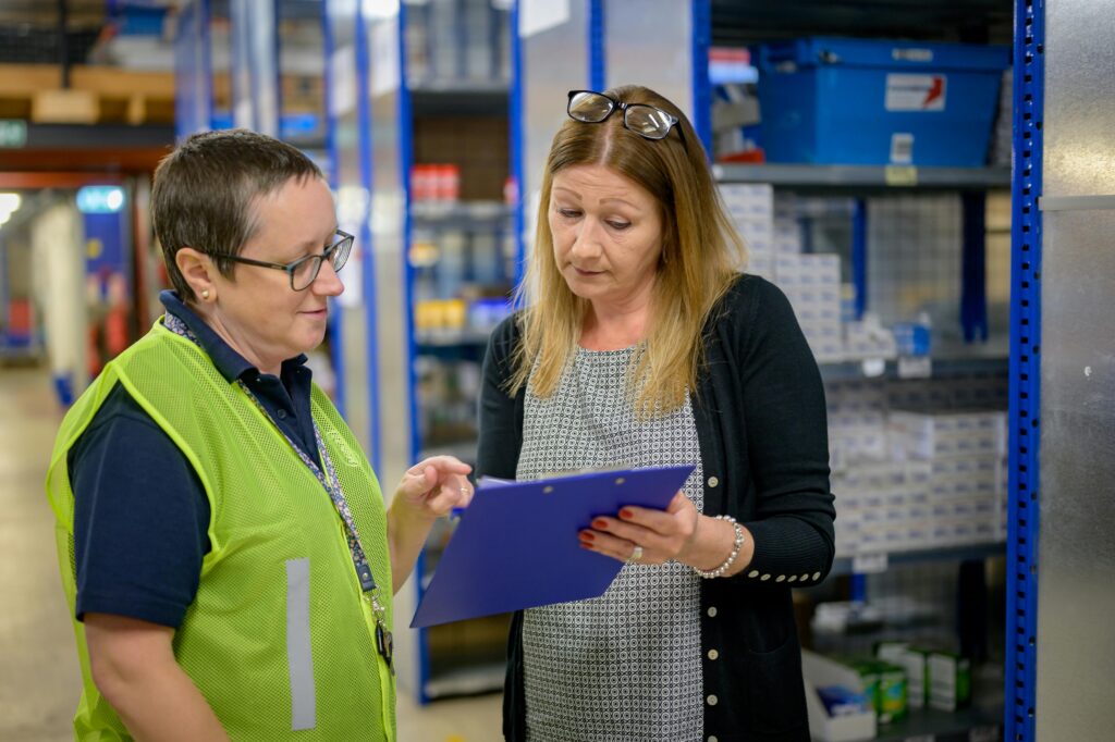 A logistics manager in a warehouse discussing tasks with a colleague, reviewing documents on a clipboard—representing hands-on coordination and operational leadership.