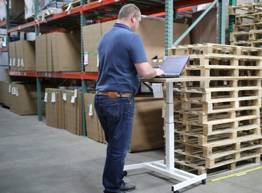 A warehouse manager working on a laptop in front of stacked pallets, highlighting inventory oversight, real-time data management, and floor-level decision-making.