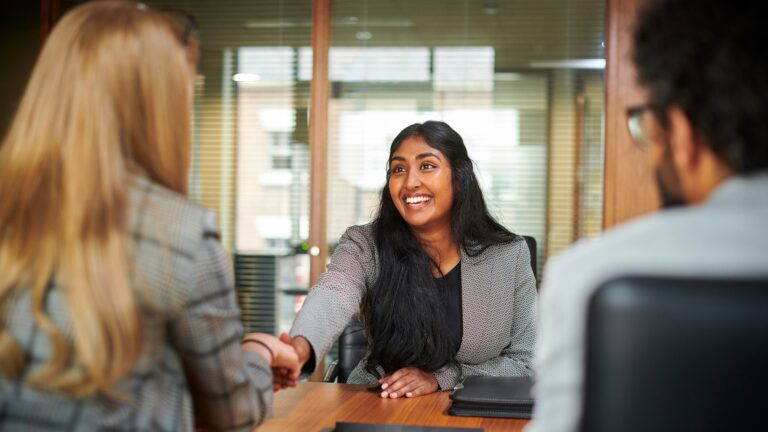 Professional young woman sitting across from two people during a job interview and shaking one of their hands.