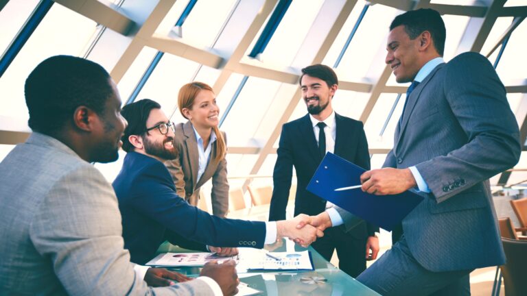 A diverse group of business professionals around a table with two people shaking hands.