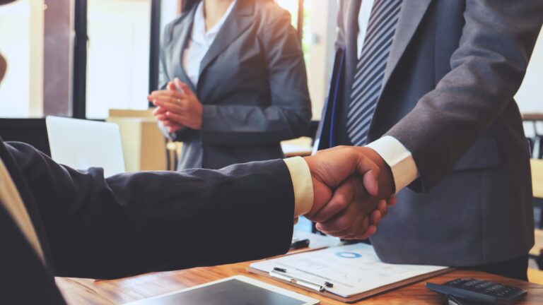 Three business professionals standing around a desk with two of them shaking hands.