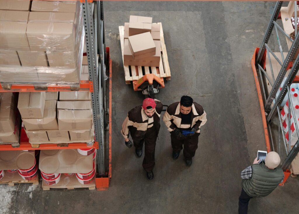 wo warehouse associates walk between shelving racks while a supervisor checks inventory on a tablet—illustrating frontline logistics teamwork and warehouse coordination.
